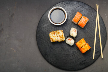 Fototapeta na wymiar Assorted rolls on a black board next to sticks, soy sauce and ginger on a dark background.