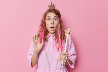 Shocked scared young woman with long hair stares impressed at camera reacts on something scarying holds magic wand wears small crown on head isolated over pink studio background. Omg concept