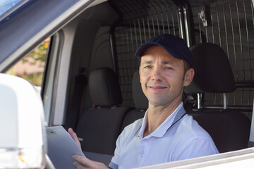 Smiling deliveryman checking documents in car. Mid adult man in white T-shirt holding pen and...