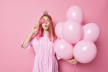 Obraz na płótnie Canvas Impressed astonished long haired woman covers eyes with heart shaped candy on stick wears dress and crown holds bunch of inflated balloons comes on birthday party isolated over pink background