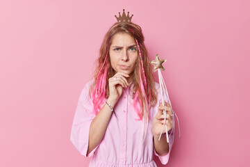 Upset discontent beautiful young woman has sulking expression raises eyebrows wears princess crown and dress holds magic wand ready to make your dreams come true isolated over pink background. - obrazy, fototapety, plakaty