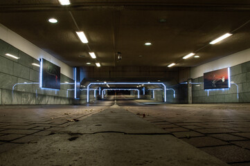 Picture of a modern underground building in the city. It is a tunnel for bikes and pedestrians. With lights above and on the sides, the lights reflecting in the walls. And paintings on the walls.