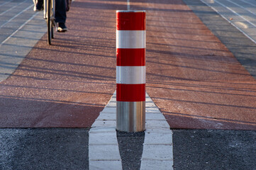 A red and white pole in the middle of a red bike path. A biker in the background. The sun is shining