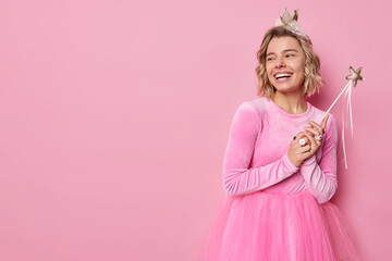 Horizontal shot of happy woman wear dress and small crown holds magic wand looks away with cheerful expression organizes party celebration for children poses against pink background copy space - obrazy, fototapety, plakaty