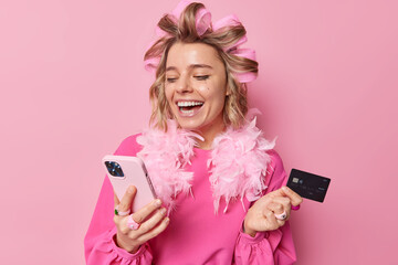 Online payment. Happy young lady with hair rollers leaked makeup laughs joyfully looks at smartphne...