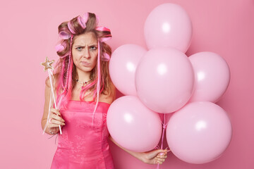 Displeased woman purses lips looks angrily doesnt like something comes on party feels frustrated poses with inflated balloons and magic wand isolated over pink background. Negative emotions.