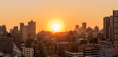A horizontal panoramic cityscape taken at sunset, against an orange sky, of the central business district of the city of Johannesburg, South Africa