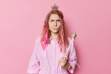 Discontent angry young woman with long hair looks gloomy wears crown and dress holds magic wand...