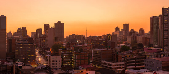 A horizontal panoramic cityscape taken after sunset against a pink and orange sky, of the central business district of the city of Johannesburg, South Africa