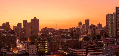 A horizontal panoramic cityscape taken after sunset, against a pink and orange sky, of the central...
