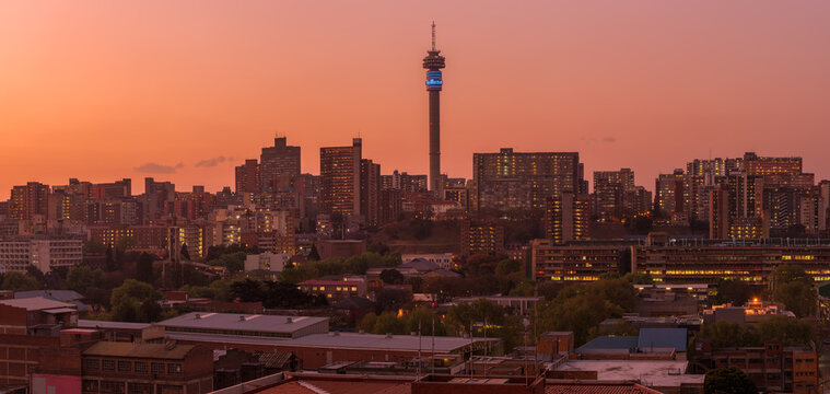 A horizontal panoramic cityscape taken after sunset against a pink and orange sky, of the central business district of the city of Johannesburg, South Africa