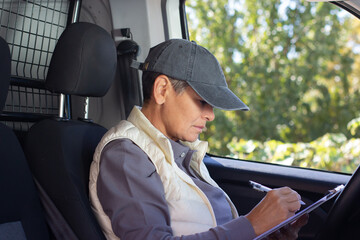 Naklejka premium Side view of deliverywoman signing papers in car. Mid adult woman in vest holding pen and documents against steering wheel. Work, delivery service, shipment concept