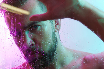 Fototapeta premium Portrait of handsome young man captured through wet glass in colorful light