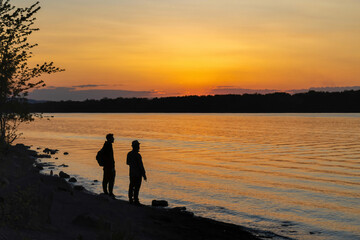 Naklejka na ściany i meble Silhouette of two men standing on a rocky shoreline by a river at sunset