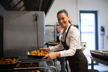 Happy female cook looking at camera and holding tray with baked pumpkin pieces in commercial kitchen