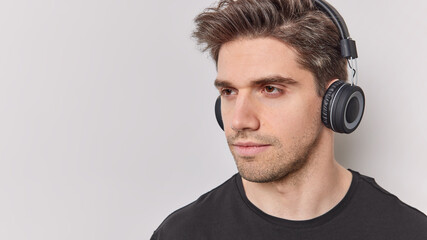Serious thoughtful man with dark hair concentrated away listens educational content via headphones poses against white background blank copy space for your advertising content. Deep thoughts