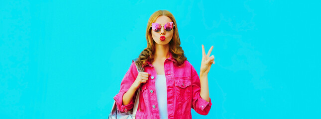 Stylish portrait of beautiful young woman blowing her red lips sending sweet air kiss wearing pink jacket, sunglasses on blue background