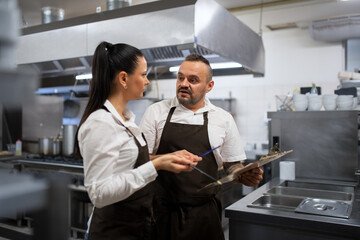 Chef and cook discussing menu indoors in restaurant kitchen.