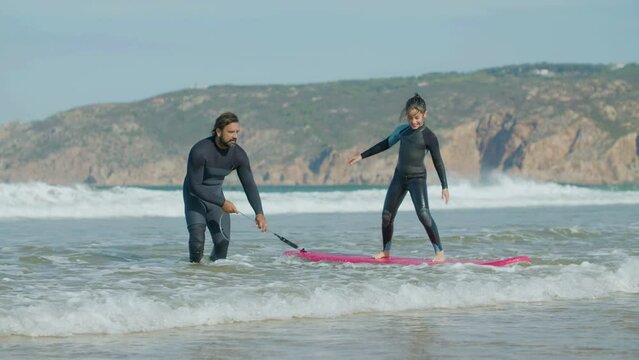 Long shot of happy girl standing on surfboard with help of coach. Slow motion of male instructor with artificial leg wearing diving suit teaching surfing in ocean. Surfing, extreme sport concept