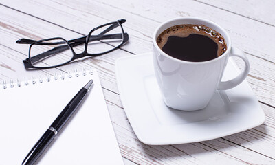 On a light wooden table is a white cup of coffee, a white notepad with a pen and black-rimmed glasses. Business concept