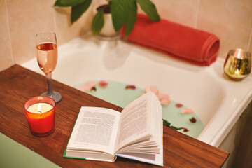 Bath with flower petals. Book, candles and glass of wine on a wood tray. Organic Spa Relaxation in...