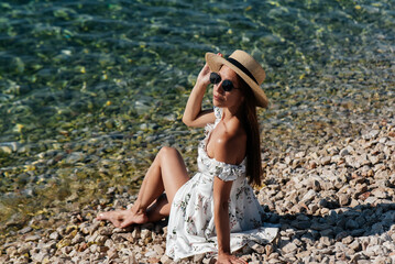 A beautiful young woman in a hat, glasses and a light dress is sitting with her back to the ocean against the background of huge rocks on a sunny day. Tourism and tourist trips.