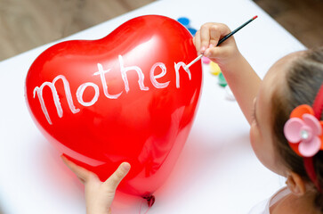 For the holiday, a 5-year-old girl writes the word mom with white paint on a heart-shaped balloon. Blurry, defocused, selective focus.