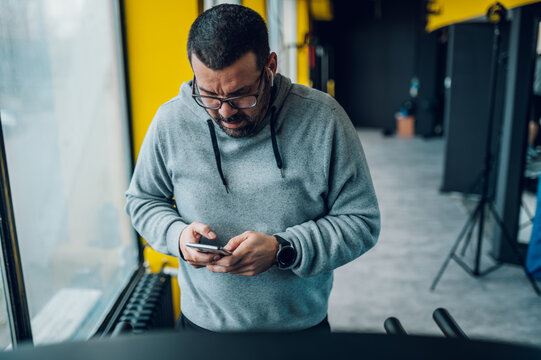 Middle Aged Man Running On Treadmill In The Gym And Using A Smartphone