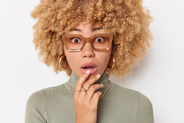 Astonished curly haired woman keeps hand on chin stares with disbelief amazed by unexpected news wears big spectacles and casual turtleneck isolated over white background. Human reactions concept