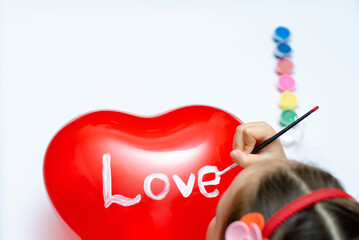 For the holiday, a 5-year-old girl writes the word love with white paint on a heart-shaped balloon. Blurry, defocused, selective focus.