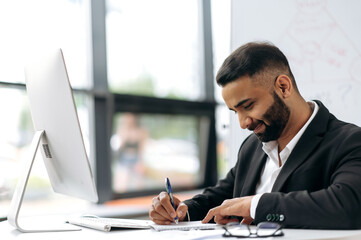 A handsome successful Indian man, financial leader, corporate manager, sits at his desk in front of a computer screen, analyzes, draws up a work plan, makes calculations, writes notes in a notebook