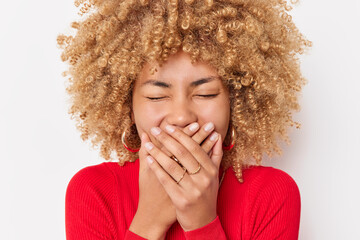 Overjoyed curly haired sincere woman laughs out joyfully covers mouth with hands tries to hide...