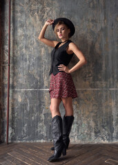 Thoughtful blonde woman model with hat in country style clothes with cowboy boots stands near concrete wall