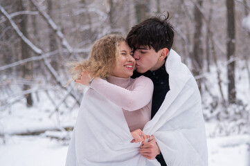 Fototapeta premium A young couple walks in the park in winter. The guy and the girl are kissing wrapped in a white blanket outdoors.
