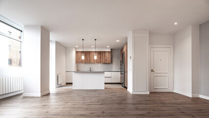 Large white and wooden kitchen with island with gray marble countertop open to an empty living room with wooden flooring