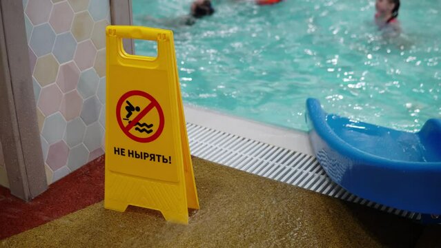 The inscription in Russian do not dive. A yellow sign stands near the pool with a warning inscription against the background of people floating in the pool