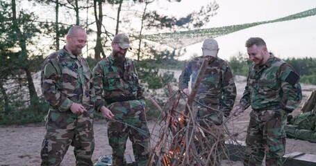 Conversation of soldiers in camouflage clothes, they are discussing in the evening by the common bonfire frying sausages for supper, men are resting after work, service, field exercises