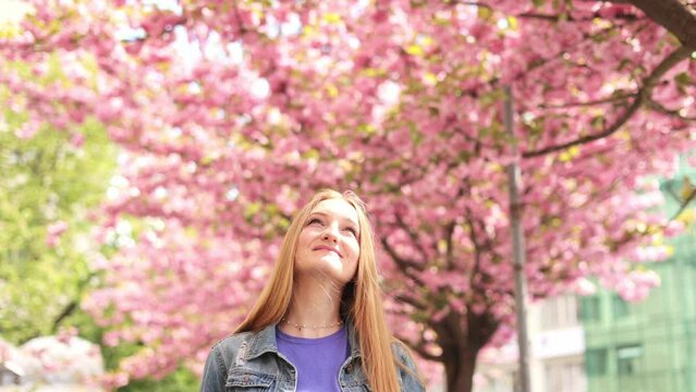 Portrait of beautiful young woman look around and smiling on blurred background in the city street. Sakura tree. Happy Caucasian beautiful female walking around Cherry blossom trees.
