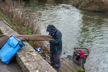 fish angler stood on a wooden jetty prepares his fishing hook bait to cast in to the flowing river 