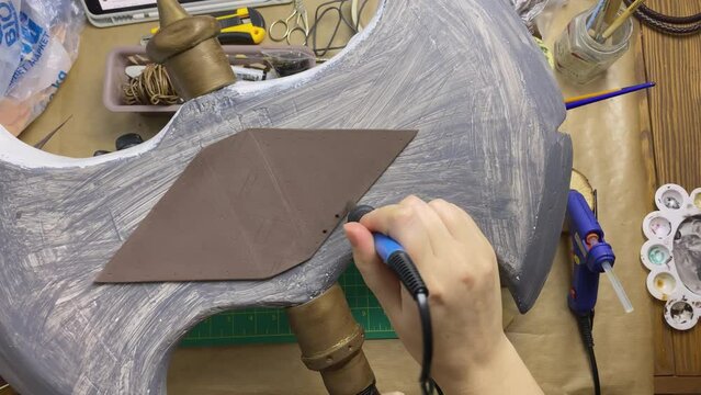 Person's hands make small holes in hatchet made of eva foam and styrofoam. Decorating with soldering iron. Celtic axe for cosplayer festival.