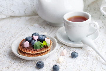 Waffles with chocolate,  blueberries, mint, marshmallow on a white lace. Romantic breakfast with white teapot, cup of tea, white spoon and fruit on waffles