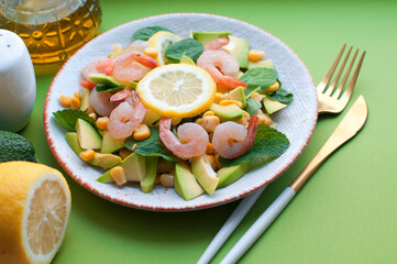 Salad with shrimp, avocado, corn and lemon with olive oil on a green background. Salad with shrimp and avocado on white plate with fork and knife