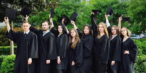 group of university or college graduates in black robes raised their hands with square caps of...