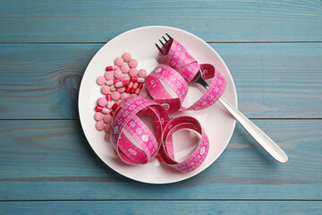 Plate with weight loss pills, measuring tape and fork on light blue wooden table, top view