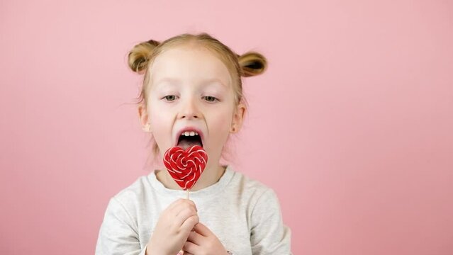 Funny little blonde girl smiling and licking red heart shape lollipop caramel on pink background. Valentines Day or Mothers Day concept