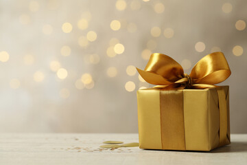 Beautiful golden gift box on light table against blurred festive lights, space for text