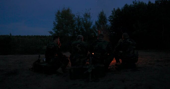 A team of men in camouflage clothing is sitting on a hill in the field at night, warming themselves by a lit campfire, frying sausages for dinner, a manly outing, relaxing after work, service, hunting