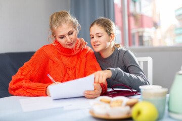 Smiling mother and her happy teenage daughter read letter from school with test results, sitting indoor
