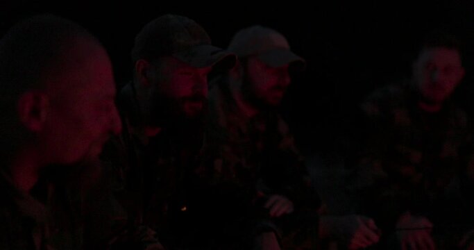 A team of men in camouflage clothing is sitting in the field at night, warming themselves by a lit campfire, frying sausages for dinner, a manly outing, relaxing after work, service, hunting