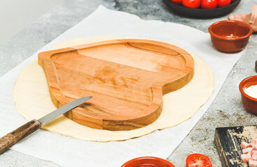 A wooden plate in the shape of a heart with a knife lie on a round thin dough .